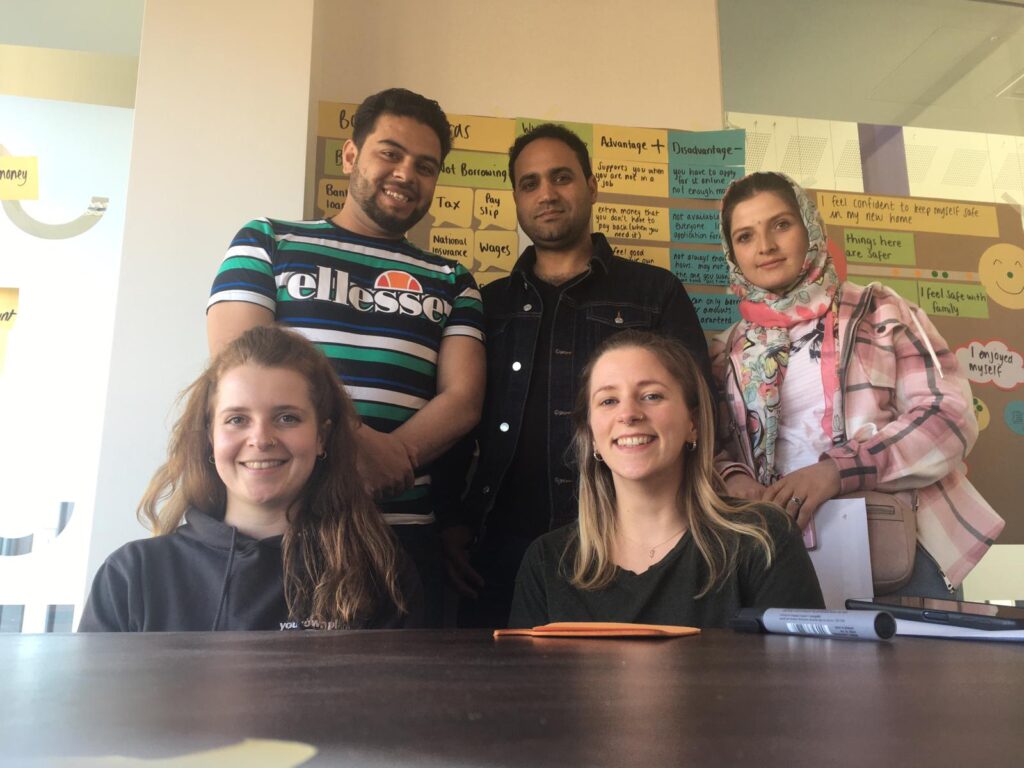 Three people standing — one is wearing a head covering, two have short black hair, and one has a beard, with two people (workshop facilitators) sitting